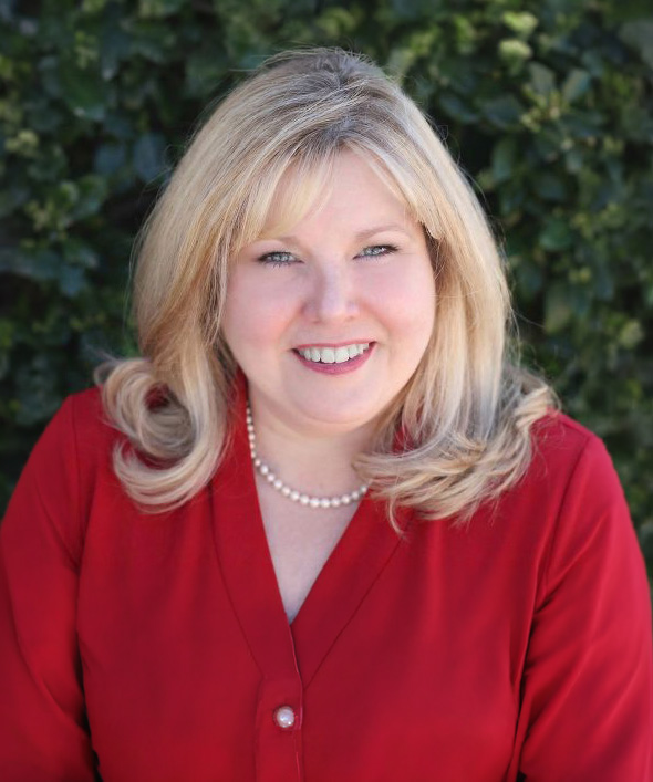 A woman with blonde hair, wearing a red blouse and a pearl necklace, smiles while standing outdoors in front of green foliage.
